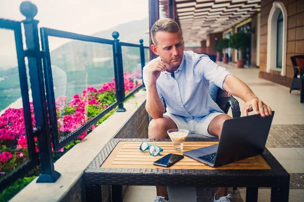 Başarılı bir erkek işadamı dizüstü bilgisayarla masada oturuyor ve tatilde. Tatil ve uzaktan kumanda. Adam terasta panoramik manzaralı bir restoranda oturuyor.