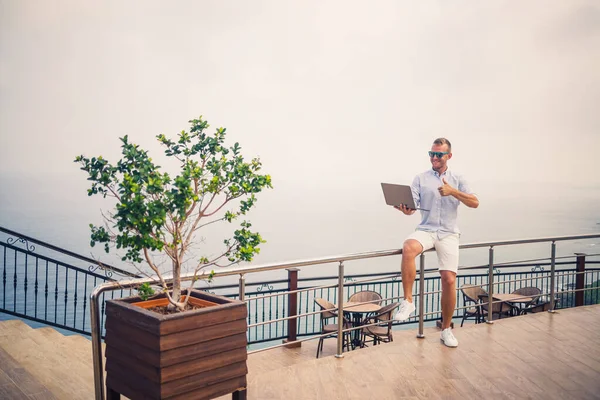 Dizüstü bilgisayarla çalışan yakışıklı ve başarılı genç iş adamı Akdeniz 'e bakıyor. O bir gömlek ve beyaz şort giyiyor. Tatilde uzaktan çalışma. Tatil kavramı