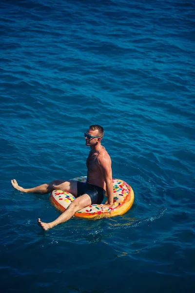 Genç bir adam güneşli bir günde şişme bir yüzükle açık denizde yüzer. Yaz tatili, tatildeki turist.