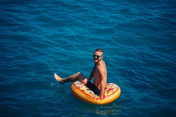 Genç bir adam mavi suyla denizde şişirilebilir bir halka üzerinde yüzüyor. Güneşli bir günde güzel bir tatil. Tatil konsepti, üst görünüm.