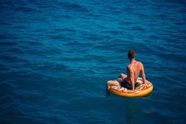 Genç bir adam mavi suyla denizde şişirilebilir bir halka üzerinde yüzüyor. Güneşli bir günde güzel bir tatil. Tatil konsepti, üst görünüm.