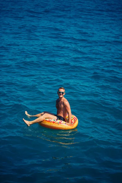 Genç bir adam mavi suyla denizde şişirilebilir bir halka üzerinde yüzüyor. Güneşli bir günde güzel bir tatil. Tatil konsepti, üst görünüm.