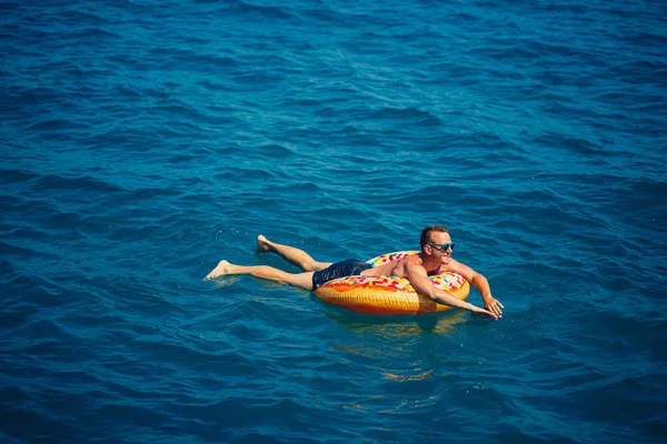 Genç bir adam mavi suyla denizde şişirilebilir bir halka üzerinde yüzüyor. Güneşli bir günde güzel bir tatil..