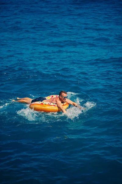 Bir adam şişme kauçuk bir çemberin üzerinde uzanır ve açık güneşli bir yaz gününde mavi denizde yüzer.