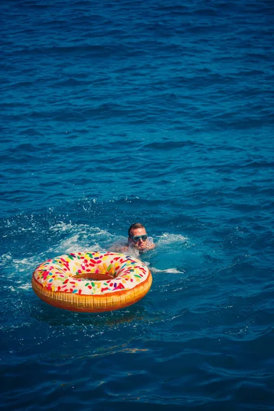 Genç bir adam mavi suyla denizde şişirilebilir bir halka üzerinde yüzüyor. Güneşli bir günde güzel bir tatil..