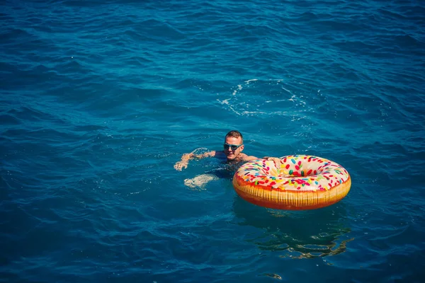 Genç adam güneşli bir günde denizde şişme bir yüzükte dinleniyor ve yüzüyor.