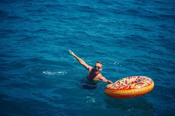 Genç adam güneşli bir günde denizde şişme bir yüzükte dinleniyor ve yüzüyor.