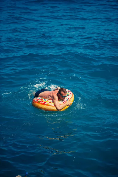 Genç adam güneşli bir günde denizde şişme bir yüzükte dinleniyor ve yüzüyor.