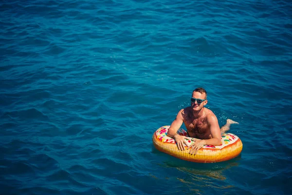 Genç adam güneşli bir günde denizde şişme bir yüzükte dinleniyor ve yüzüyor.