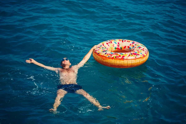 Genç adam güneşli bir günde denizde şişme bir yüzükte dinleniyor ve yüzüyor.