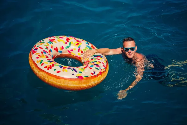 Genç adam güneşli bir günde denizde şişme bir yüzükte dinleniyor ve yüzüyor.