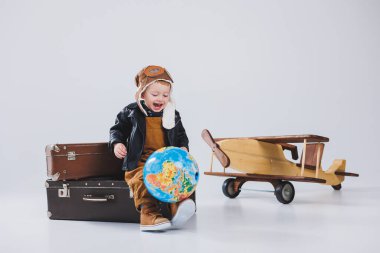 Deri ceketli ve pilot şapkalı bir çocuk, tahta bir uçak, bir küre, kahverengi bavullar. Çocukların tahta oyuncakları.