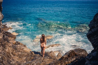 Deniz fırtınası sırasında kayalık bir sahilde uzun saçlı, mayo giymiş seksi bir kadın büyük kayalıklarda yürüyor. Arkadan bak. Kadın mayo adası tropik manzara egzotik yürüyüşü