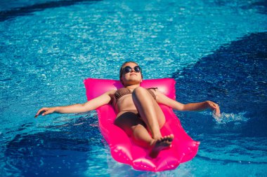 Havuzdaki pembe şişme yatakta mayo giymiş seksi bir kadın yatıyor. Sıcak ve güneşli bir yaz gününde havuz kenarında rahatla. Tatil kavramı