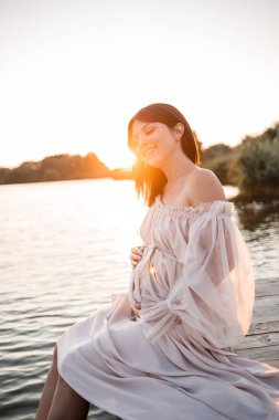 Nehrin kenarında güzel bir gün batımı olan narin elbiseli hamile bir kadın. Güzellik, sağlık ve nazik annelik. Mutlu annelik