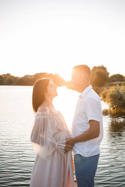 妊娠中の女性と彼女の夫は桟橋に立って お互いを見て 美しい川と日没 将来の両親は屋外で時間を過ごす 美しい若い母親 — ストック写真