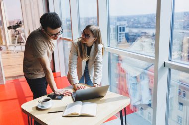 İki meslektaş, bir erkek ve bir kadın yönetici, modern bir ofiste dizüstü bilgisayarla bir masanın yanında duruyor ve yeni bir projeyi tartışıyorlar. Gözlüklü iki iş ortağı yeni bir iş hakkında konuşuyor..