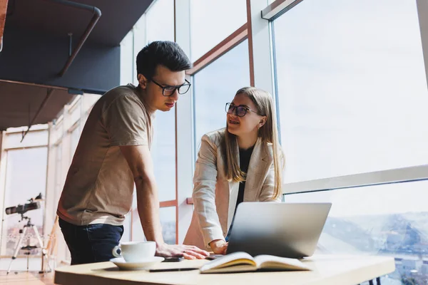 İki meslektaş, bir erkek ve bir kadın yönetici, modern bir ofiste dizüstü bilgisayarla bir masanın yanında duruyor ve yeni bir projeyi tartışıyorlar. Gözlüklü iki iş ortağı yeni bir iş hakkında konuşuyor..