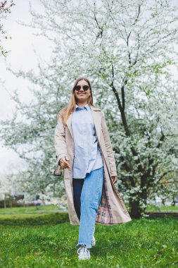 Prag 'daki tepedeki tepede, doğal parktaki çiçek açan elma ağacının altında yürüyen arka plan kızı. Şapkalı ve kahverengi ceketli zarif genç bayan güneşli bahar gününde çiçek açan bahçenin tadını çıkarıyor..