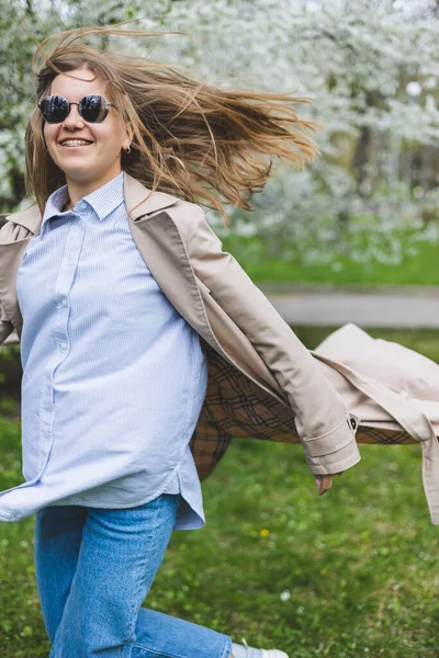 Sarı saçlı genç bir kadın çiçek açan bir bahar bahçesinin tadını çıkarıyor. Seyahat, bahar tatili. Modaya uygun. Güneş gözlüklü ve bej renkli trençkotlu bir kadın çiçek parkında koşuyor. Seçim odağı
