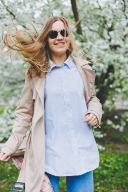 Sarı saçlı genç bir kadın çiçek açan bir bahar bahçesinin tadını çıkarıyor. Seyahat, bahar tatili. Modaya uygun. Güneş gözlüklü ve bej renkli trençkotlu bir kadın çiçek parkında koşuyor. Seçim odağı