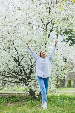Bahar bahçesinde elinde gözlük olan beyaz gömlekli ve kot pantolonlu bir kadın..