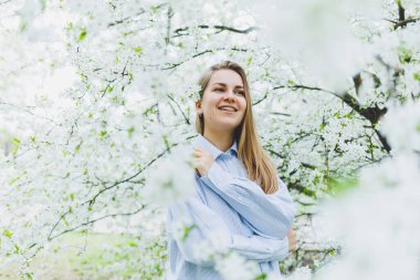 Genç ve güzel bir kadın çiçek açan bahar ağacının yanında durmaktan hoşlanıyor. Bej şapkalı ve beyaz elbiseli bir kız çiçek açan elma ağaçlarının arasında gülümsüyor. Bahar mevsimi konsepti