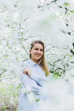 Genç ve güzel bir kadın çiçek açan bahar ağacının yanında durmaktan hoşlanıyor. Bej şapkalı ve beyaz elbiseli bir kız çiçek açan elma ağaçlarının arasında gülümsüyor. Bahar mevsimi konsepti