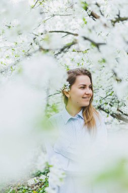 Genç ve güzel bir kadın çiçek açan bahar ağacının yanında durmaktan hoşlanıyor. Bej şapkalı ve beyaz elbiseli bir kız çiçek açan elma ağaçlarının arasında gülümsüyor. Bahar mevsimi konsepti