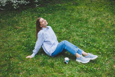 Kot ceketli genç hippi kadın parktaki yeşil çimlerde dinleniyor, kahve içiyor, dışarıda dinleniyor. Yaşam tarzı, kahve molası. Tek kullanımlık kahveyi tutan kadın ellerine odaklan..