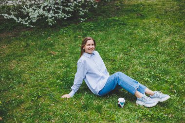 Kot ceketli genç hippi kadın parktaki yeşil çimlerde dinleniyor, kahve içiyor, dışarıda dinleniyor. Yaşam tarzı, kahve molası. Tek kullanımlık kahveyi tutan kadın ellerine odaklan..