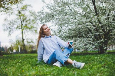 Kot ceketli genç hippi kadın parktaki yeşil çimlerde dinleniyor, kahve içiyor, dışarıda dinleniyor. Yaşam tarzı, kahve molası. Tek kullanımlık kahveyi tutan kadın ellerine odaklan..