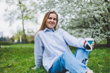 Kot ceketli genç hippi kadın parktaki yeşil çimlerde dinleniyor, kahve içiyor, dışarıda dinleniyor. Yaşam tarzı, kahve molası. Tek kullanımlık kahveyi tutan kadın ellerine odaklan..