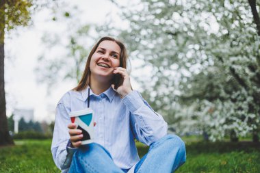 Parkın bahçesindeki yeşil çimlerde oturan kadının en iyi görüntüsü. Elinde akıllı telefon, kulaklık, tablet, kitap ve kahve var.