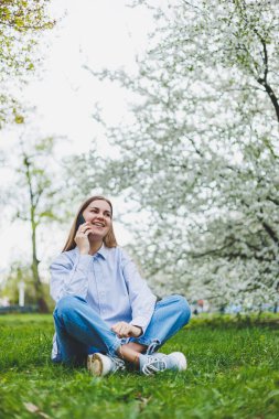Şehir caddesinde akıllı telefon kullanarak yürüyen mutlu bir kadın. Güneş gözlüklü iş kadını dışarıda, ağacın altında cep telefonu kullanıyor. Kız mesaj yazıyor, sohbet ediyor, web sayfası kayıyor. Kameraya bakın.