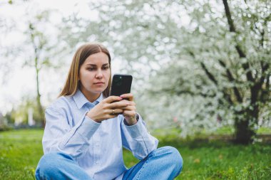 Happy woman walking using smart phone in a city street. Business woman with sunglasses uses mobile phone outdoors under blooming tree, girl typing message, chatting, scrolling web page, look at camera