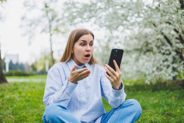 Genç duygusal Asyalı kadın cep telefonu kullanıyor internetten video izliyor, çimlerin üzerinde oturan haberleri yetiştiriyor. İnternetten heyecanlı kadın alışverişi