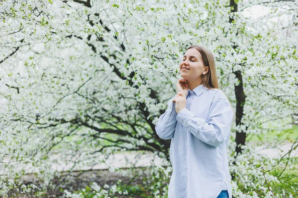 若いきれいな女性は春の木の近くに立って楽しんでいます ベージュの帽子と白いドレスを着た女の子がリンゴの木の間で微笑む 春の季節のコンセプト — ストック写真