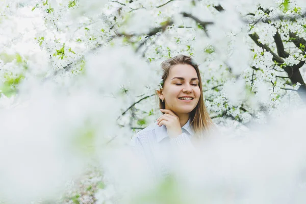 Genç ve güzel bir kadın çiçek açan bahar ağacının yanında durmaktan hoşlanıyor. Bej şapkalı ve beyaz elbiseli bir kız çiçek açan elma ağaçlarının arasında gülümsüyor. Bahar mevsimi konsepti