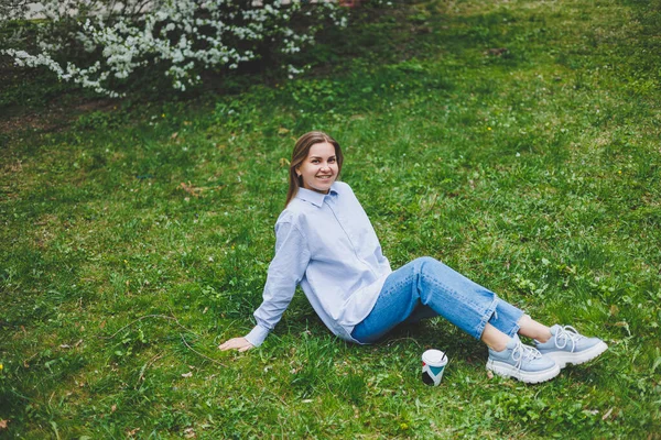 Kot ceketli genç hippi kadın parktaki yeşil çimlerde dinleniyor, kahve içiyor, dışarıda dinleniyor. Yaşam tarzı, kahve molası. Tek kullanımlık kahveyi tutan kadın ellerine odaklan..