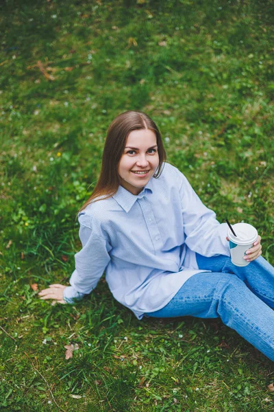 Kot ceketli genç hippi kadın parktaki yeşil çimlerde dinleniyor, kahve içiyor, dışarıda dinleniyor. Yaşam tarzı, kahve molası. Tek kullanımlık kahveyi tutan kadın ellerine odaklan..