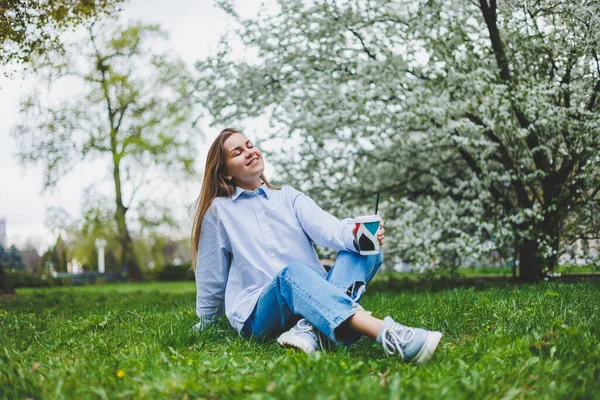 デニムジャケットの若いヒップスター女性が公園の緑の芝生の上で休んでいる コーヒーを飲んで 屋外でリラックスしています ライフスタイルコーヒー休憩 コーヒーの使い捨てカップを保持する女性の手に選択的な焦点 — ストック写真