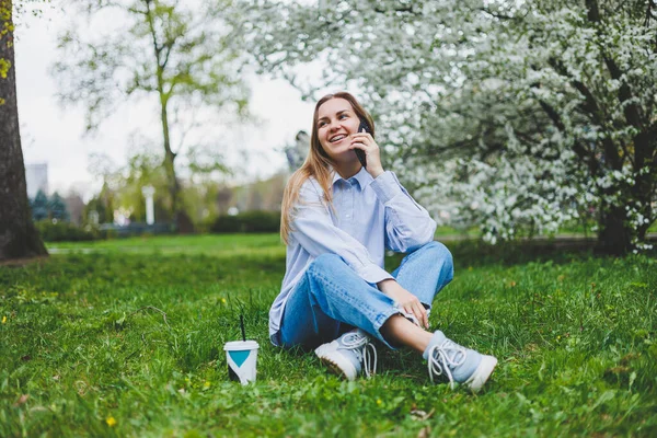 Parkın bahçesindeki yeşil çimlerde oturan kadının en iyi görüntüsü. Elinde akıllı telefon, kulaklık, tablet, kitap ve kahve var.