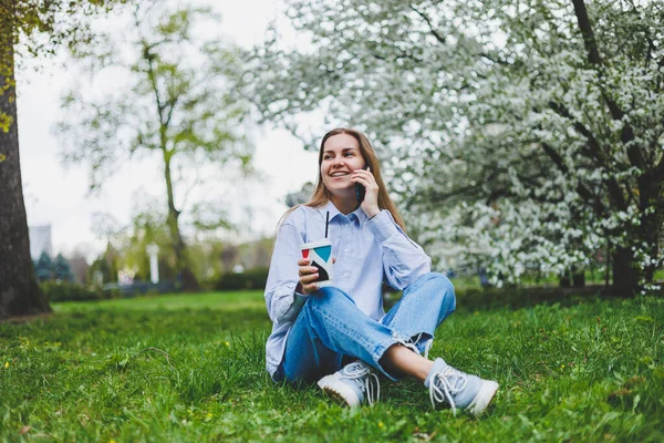 Parkın bahçesindeki yeşil çimlerde oturan kadının en iyi görüntüsü. Elinde akıllı telefon, kulaklık, tablet, kitap ve kahve var.
