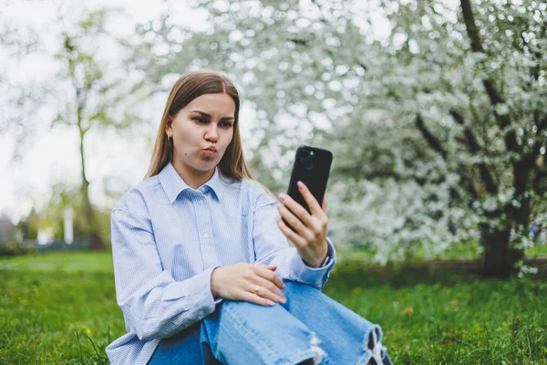 Happy woman walking using smart phone in a city street. Business woman with sunglasses uses mobile phone outdoors under blooming tree, girl typing message, chatting, scrolling web page, look at camera