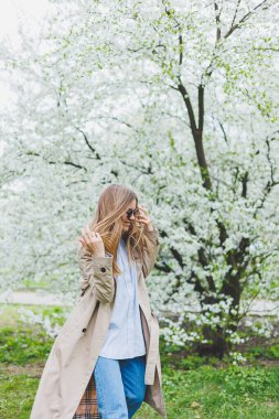 Sarı saçlı genç bir kadın çiçek açan bir bahar bahçesinin tadını çıkarıyor. Seyahat, bahar tatili. Modaya uygun. Güneş gözlüklü ve bej renkli trençkotlu bir kadın çiçek parkında koşuyor. Seçim odağı