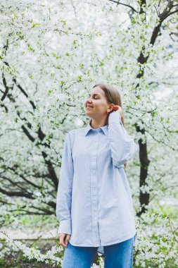 Genç ve güzel bir kadın çiçek açan bahar ağacının yanında durmaktan hoşlanıyor. Bej şapkalı ve beyaz elbiseli bir kız çiçek açan elma ağaçlarının arasında gülümsüyor. Bahar mevsimi konsepti