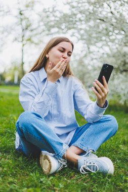 Happy woman walking using smart phone in a city street. Business woman with sunglasses uses mobile phone outdoors under blooming tree, girl typing message, chatting, scrolling web page, look at camera