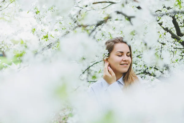 若いきれいな女性は春の木の近くに立って楽しんでいます ベージュの帽子と白いドレスを着た女の子がリンゴの木の間で微笑む 春の季節のコンセプト — ストック写真