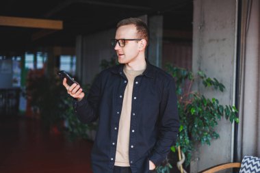 Optik gözlüklü mutlu bir adamın yarım boy portresi modern cep telefonunu tutuyor ve kameraya gülümsüyor, dijital akıllı telefonlu neşeli blogcu birlikte çalışma alanında poz veriyor.